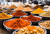 Rows of mix colored powdered spices and seeds in trays at local market stall over blurred busy market background