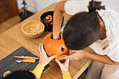 From above of crop anonymous female friends looking down while standing at wooden table with Halloween tools bowls and carving mouth in daylight