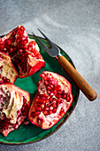 From above fresh pomegranate pieces with vibrant red seeds on a green ceramic plate set against a textured grey concrete background