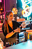 Confident young female barkeeper in black uniform pouring alcohol in jigger from glass bottle at illuminated bar counter