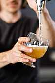 Anonymous young unshaven male looking down while standing with transparent glass in hand opening tap and pouring beer in daylight against blurred interior