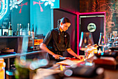 Side view of Focused young female chef with braids in black uniform with focus cutting fish with knife on chopping board while preparing meal at illuminated counter in bistro