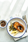 Top view of well-prepared grilled trout steak with capers and lemon served on a white plate with a blue rim placed on striped napkin, cutlery and bowl with capers