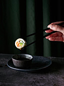 Sushi roll held by anonymous person using chopsticks with a droplet of sauce ready to fall in bowl placed on table against green curtains