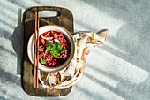 Top view of bowl with beetroot soup with onion, coriander and noodles in Asian style served on bowl and chopsticks on wooden cutting board near napkin against gray surface in daylight