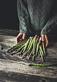 Unrecognizable person in gray sweater standing at wooden table with fresh asparagus stems