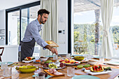 Side view of Bearded male cook in apron putting pineapple slices with tongs on plate while serving dishes for breakfast in modern dining room