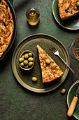 Draufsicht auf eine Scheibe gebackenes italienisches Focaccia-Brot mit grünen Oliven in einem Teller auf einem Esstisch mit Getränk auf grünem Hintergrund