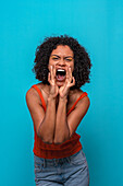Joyful African American female model with Afro hairstyle wearing casual clothes shouting against blue background