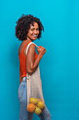 Side view of delighted African American female model with Afro hairstyle in casual clothes carrying bag of fruit on shoulder against blue background