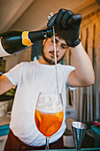 Focused young professional barman pouring sparkling wine into orange drink while preparing refreshing cocktail in bar