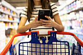 Crop anonymous female customer in casual clothes using smartphone while shopping alone at supermarket