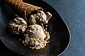 Black ceramic plate with a cone of chocolate ice cream accompanied by two scoops of ice cream with peanuts and walnuts on a dark concrete background