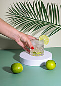 Anonyme Frau mit rotem Nagellack, die ein Glas mit einem Cocktailgetränk mit Eis und einer Zitrusscheibe auf einem weißen Tisch mit Zitronen neben Blättern abstellt