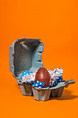 Close-up of some blue and red easter eggs inside a gray cardboard box on an orange background