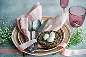 Elegant gedeckter Ostertisch mit pastellfarbenen Servietten, Tellern und dekorativen Eiern in einem Zweignest, ergänzt durch Blumen und Bänder
