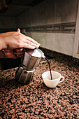 Anonyme Hand mit türkisfarbenem Armband gießt frisch gebrühten Kaffee aus einer Espressomaschine auf dem Herd in eine weiße Tasse vor einem modernen Küchenhintergrund