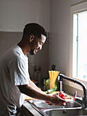 Side view of young ethnic African American male in casual t shirt washing fresh bell pepper in sink with water stream while cooking healthy salad in kitchen