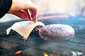 Hand placing cheese slice on burger patty over a flaming grill