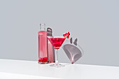 Glass filled with red pomegranate cocktail and adorned with pomegranate seeds, stands beside a transparent bottle of the same vibrant drink, both casting soft shadows on a calm gray background