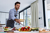 Low angle of bearded male private chef in apron serving yummy pineapple slices on plate with strawberries while preparing breakfast in dining room