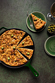 Top view of flat leavened oven-baked Italian focaccia bread in pan served with fresh green olives in bowls on green background surface