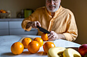 Crop anonymous senior man in casual clothes cutting fresh organic oranges with knife on table while sitting in kitchen at home