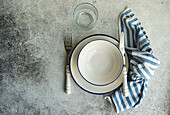 Draufsicht auf einen minimalistisch-rustikal gedeckten Tisch mit weißen Tellern, Besteck, Glas und gestreifter Serviette auf grauer Fläche
