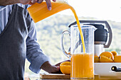 Anonymous male chef in apron standing at kitchen table with appliance and pouring squeezed fresh orange juice into jug during healthy breakfast preparation against blurred background in daylight