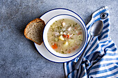 Draufsicht auf eine Schüssel mit Gemüse- und Hühnersuppe und einer Scheibe Brot auf einer grauen Fläche mit einer blau-weiß gestreiften Serviette und einem Löffel an der Seite