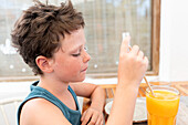 Side view of thoughtful cute boy drinking mango juice from glass and looking away seriously while sitting at table in cafe during summer vacation