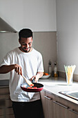 Positive young ethnic African American male in casual clothes preparing delicious dish while frying tomatoes on pan and mixing ingredients with wooden spatula during cooking process in kitchen