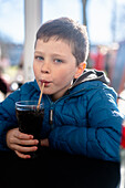 A young boy in a blue jacket looking at camera while sipping a dark soda through a straw at a table