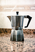 A vintage silver moka pot stands on a granite countertop illuminated by soft natural light