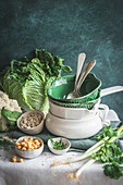 From above decorated table with various dishware ceramic bowls and spoons with fresh cauliflower, cabbage, legumes and herbs placed on table against green background