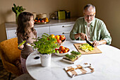 Senior man in casuals cutting lettuce with knife on chopping board while preparing salad with granddaughter on table at home