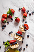 From above of fresh strawberries and blueberries with sliced banana placed in cups and on white marble table bright room