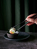 Crop hand of anonymous person using chopsticks to elegantly hold a sushi roll above a small bowl of soy sauce against dark green curtains