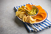 Teller mit verschiedenen Chips, darunter Kartoffel-, Paprika- und Käsesorten, serviert mit einer Schüssel frischer Avocado-Guacamole auf einer gestreiften Serviette