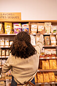 Back view of unrecognizable seller with curly hair in uniform arranging pack with various products on shelves during work in supermarket