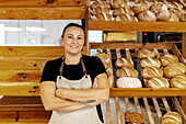 Porträt einer selbstbewussten Verkäuferin in Schürze mit verschränkten Armen, die neben frisch gebackenen Broten auf Holzregalen in einer Bäckerei steht