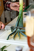 High angle of anonymous chef's hands carefully preparing leeks on a white cutting board, with the green of the leeks standing out against the kitchen's neutral tones