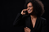 Positive Hispanic young female in outfit smiling and looking away while talking on mobile phone while standing on black background