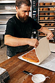 Serious bearded mature male chef with concentration joining the edges of stuffed dough on wooden table while making delicious pastries in kitchen at bakery