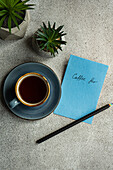 Draufsicht auf eine Tasse Kaffee neben Topfpflanzen und einer Notiz mit Bleistift auf einem grauen Tisch