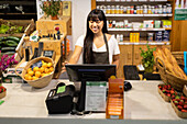 Happy young Asian female seller in apron smiling and using computer while standing at counter with various products during work in supermarket