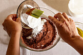Top view of Hands of crop anonymous person carefully pour sifted flour into a bowl with cocoa powder for a baking recipe against a white background