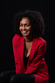 Smiling young Hispanic female model with Afro hairstyle in red coat looking at camera while sitting against black background