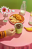 Unrecognizable person drinking hot aromatic coffee in cup placed on table with delicious croissants and banana decorated by flowers in vase