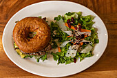Draufsicht auf einen leckeren Burger mit Rindfleisch, Käse, frischem Salat und Karottensprossen auf einem Teller auf einer hölzernen Oberfläche im Tageslicht
